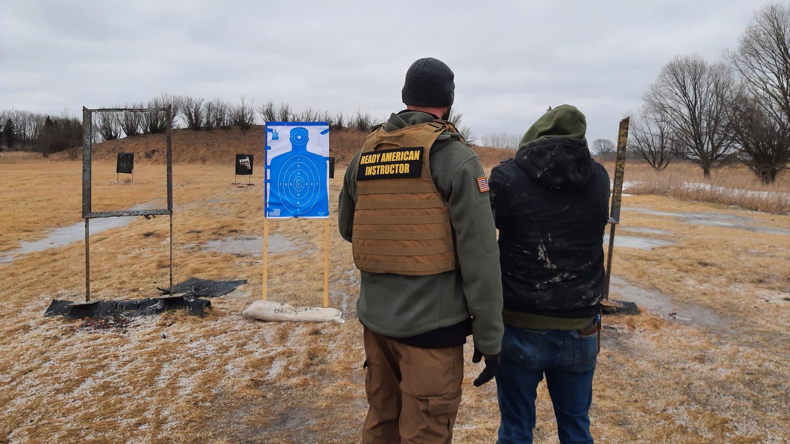 Ryan, from behind in a Ready American Instructor vest, coaching a student at the outdoor range.